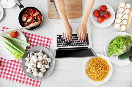 A food blogger typing on her laptop in the kitchen next to assorted ingredients.